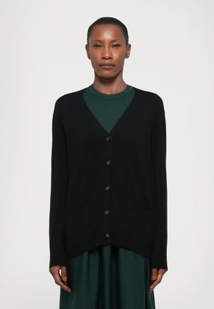 Cárdigan negro con escote en pico, bolsillos frontales y cinco botones. Textura suave, mangas largas y corte relajado sobre una blusa verde.