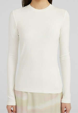 Persoon draagt een nauwsluitend, wit shirt met lange mouwen en ribbelpatroon, gecombineerd met een lichtgekleurde rok, staand tegen een effen achtergrond.