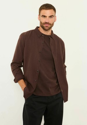 Mann mit kurzen braunen Haaren und Bart, trägt ein dunkelbraunes Hemd mit Knöpfen über einem passenden T-Shirt und schwarze Hose, Hände in den Taschen, neutraler Hintergrund.
