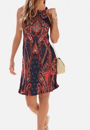 Frau trägt ärmelloses Neckholderkleid mit rotem und marineblauem Paisley-Muster, hält eine kleine geflochtene Handtasche und trägt Sandalen.