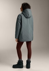 Žena koja nosi tirkiznu prošivenu jaknu s kapuljačom, vinske tajice, sive čarape i crne zimske čizme s vezicama stoji ispred jednobojne pozadine.