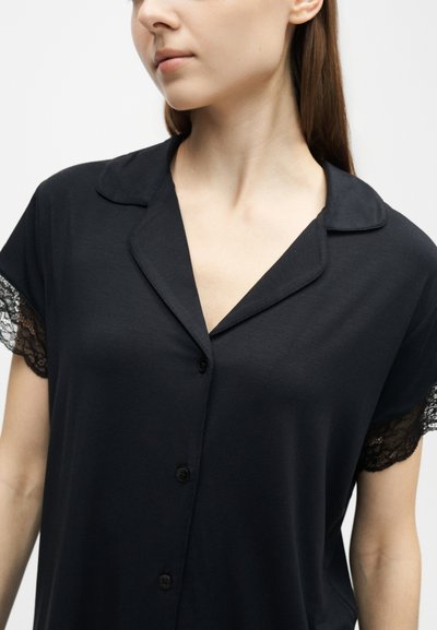 Femme portant une chemise noire boutonnée avec des manches courtes bordées de dentelle et un col cranté, légèrement tournée vers la gauche.