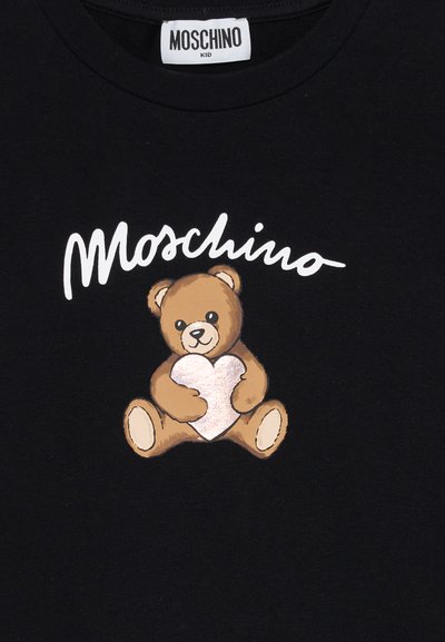 Fekete póló fehér "Moschino" felirattal egy barna mackó előtt, aki egy fényes rózsaszín szívet tart a kezében.