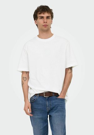 Mladi muškarac s kratkom smeđom kosom nosi običnu bijelu majicu, plave traperice i smeđi remen, stoji s jednom rukom u džepu.