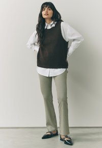 Frau steht mit den Händen in den Hüften, trägt ein weißes Hemd, einen dunkelbraunen ärmellosen Pullover, karierte Hosen und schwarze spitze Ballerinas.