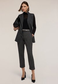 Gilet grigio con texture su una maglietta in rete nera, abbinato a pantaloni a vita alta con risvolto e tacchi a spillo neri in vernice. Gli accessori includono una borsa nera.
