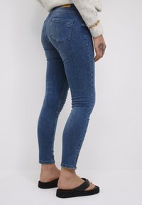 Karcsú kék farmernadrág, közepes mosással, öt zsebes dizájnnal és bokáig érő cipzárokkal, fekete szandálokkal párosítva.