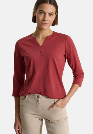 Femme aux cheveux bruns courts portant un haut rouge à manches trois-quarts et un pantalon beige clair, debout avec une main dans la poche.
