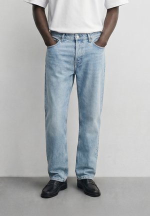 Mand iført lyseblå jeans med lige ben, med hænderne i lommerne og sorte loafers, stående mod en ensartet lys grå baggrund.