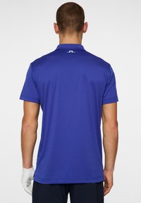 Short-sleeve polo shirt in solid electric blue fabric. Features a collar, subtle texture, and a small white logo on the upper back.