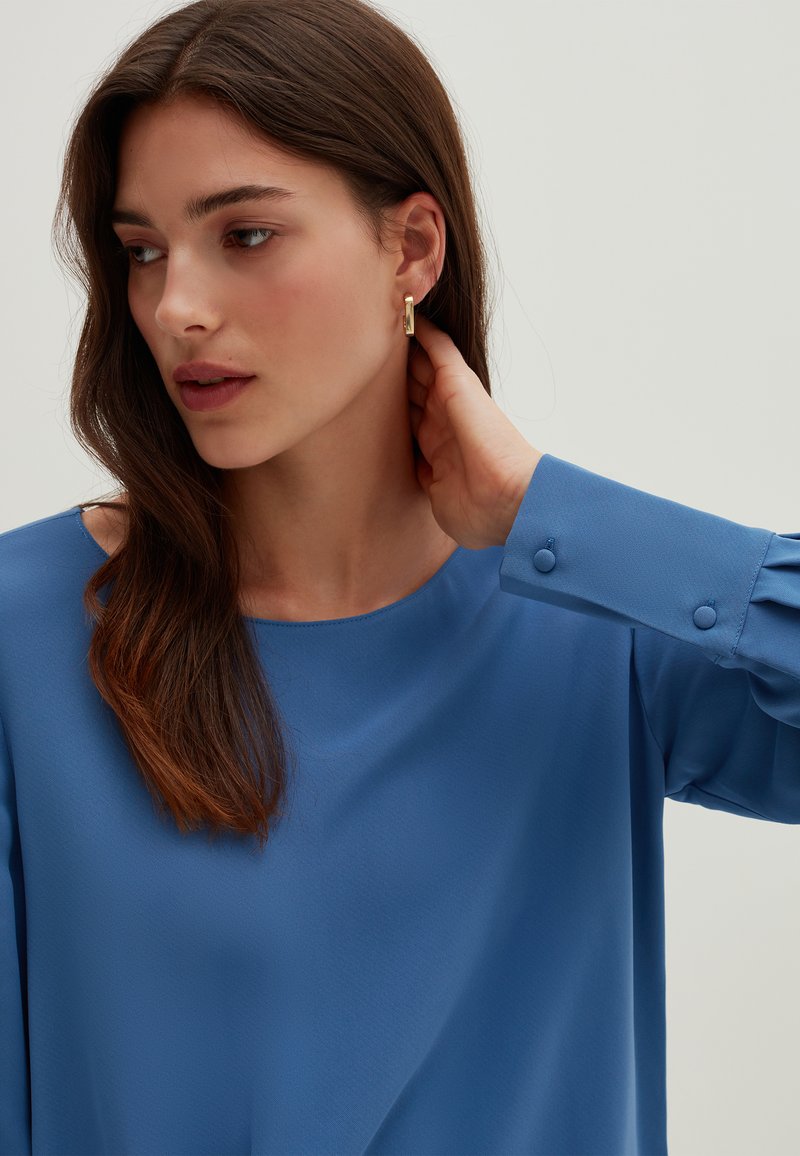 Blusa blu con scollo rotondo, maniche lunghe con dettagli in bottone e tessuto leggero. I capelli sono lunghi e ondulati. Sono visibili orecchini dorati.