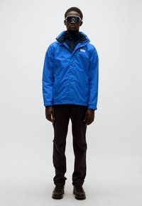 Μπλε αδιάβροχο μπουφάν με ψηλό γιακά και μαύρο εσωτερικό, συνδυασμένο με μαύρο παντελόνι και στιβαρά μαύρα παπούτσια· το μοντέλο φορά αντανακλαστικά γυαλιά ηλίου.