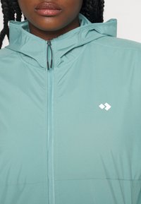Gros plan d'une personne portant une veste zippée bleu clair avec un petit logo blanc sur la poitrine et une capuche dans le dos.