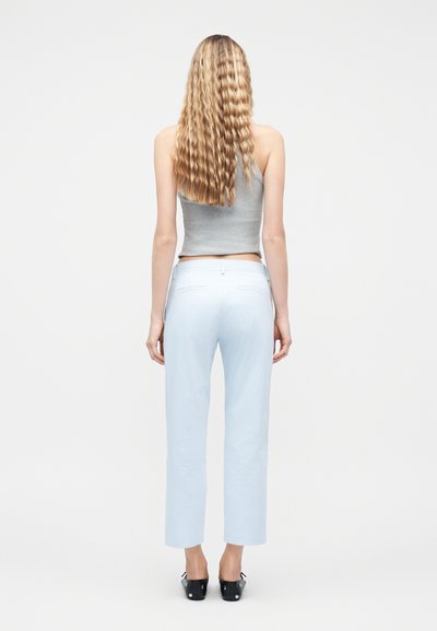 Γυναίκα με μακριά κυματιστά ξανθά μαλλιά, φορώντας γκρι αμάνικο τοπ, ανοιχτό μπλε παντελόνι ψηλόμεσο και μαύρα παπούτσια, στραμμένη προς τα πίσω σε λευκό φόντο.