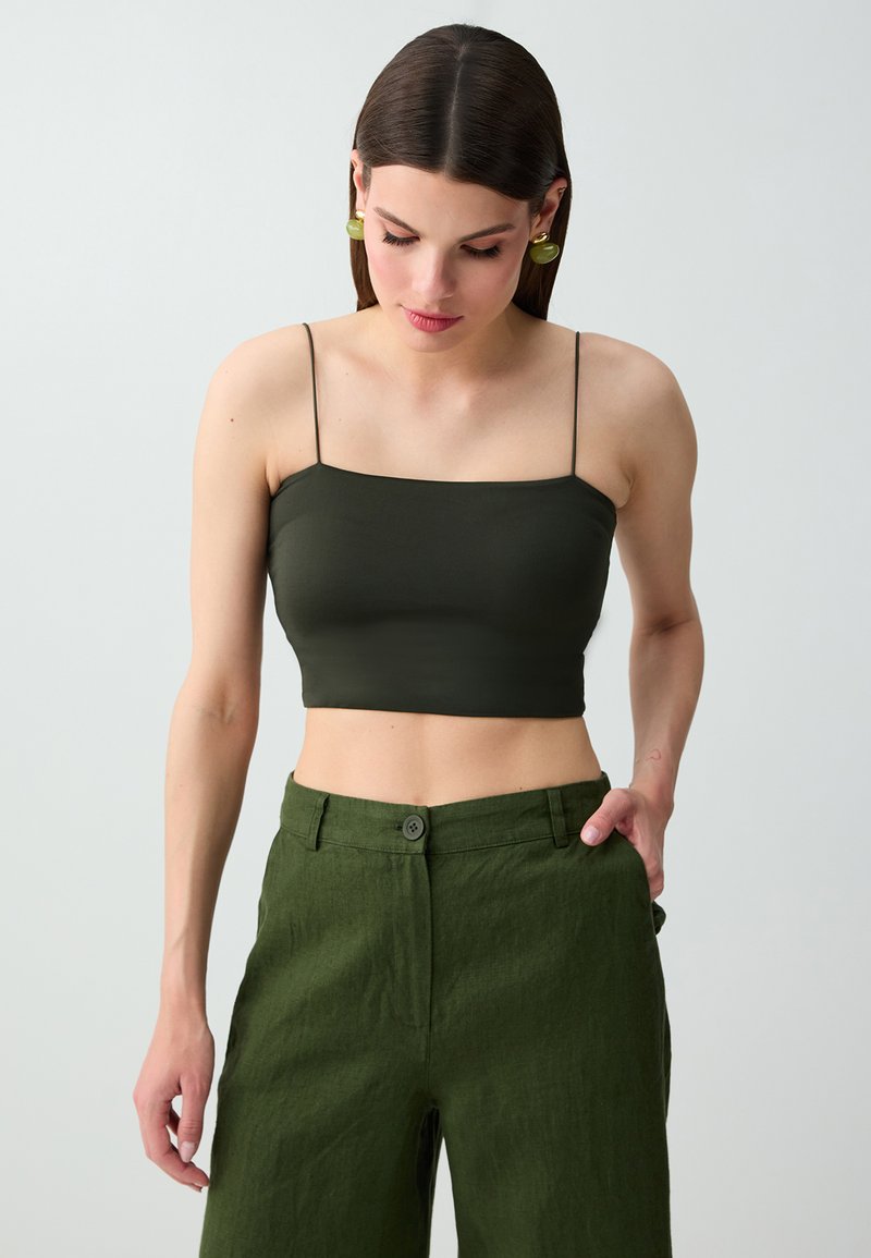 Femme portant un débardeur court vert olive foncé à fines bretelles et un pantalon vert ample taille haute, debout avec une main dans la poche.