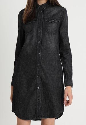 Femme portant une robe chemise en jean noir à manches longues, avec des boutons sur le devant et deux poches poitrine, debout devant un fond clair et uni.
