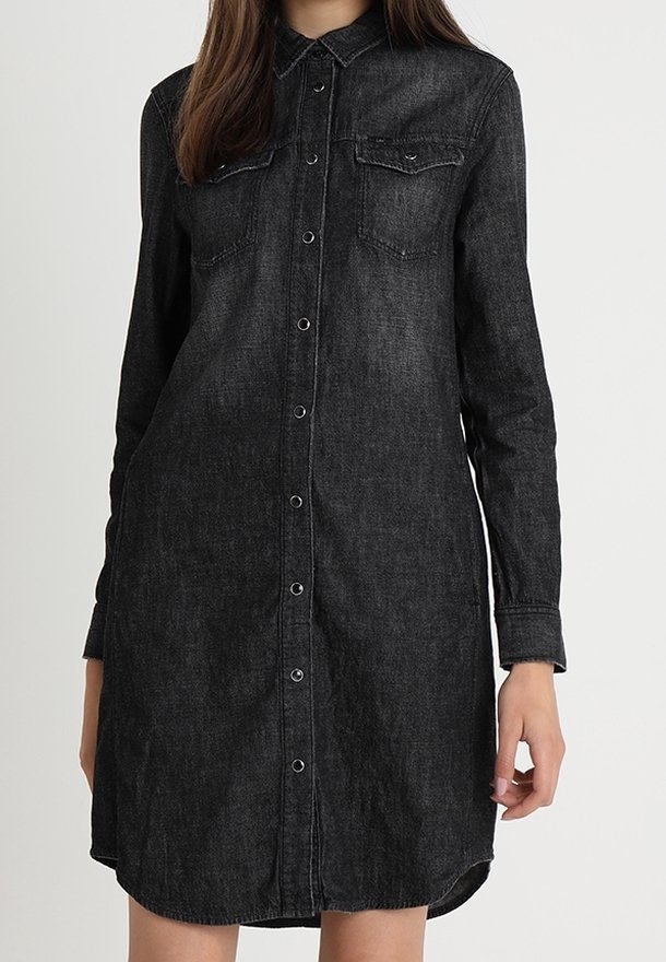 Femme portant une robe chemise en jean noir à manches longues, avec des boutons sur le devant et deux poches poitrine, debout devant un fond clair et uni.