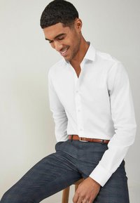 Camisa blanca de botones, de manga larga, con un bolsillo, combinada con pantalones de cuadros oscuros y un cinturón marrón. Tejido suave y diseño ajustado.