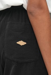 Fekete kordbársony nadrág oldalzsebbel, amelyen egy bézs bőr folt található, stilizált logóval debütálva. Az anyagnak bordázott textúrája van.