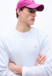 Νέος άντρας με ανοιχτό μπλε μακρυμάνικο πουκάμισο με το λογότυπο Calvin Klein, φοράει φωτεινό ροζ καπέλο, κοιτάζοντας προς τα πλάγια με σταυρωμένα τα χέρια.