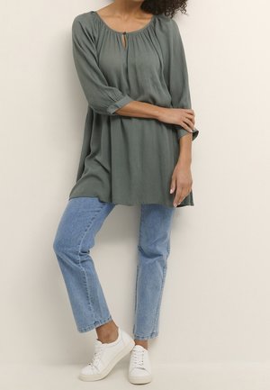Tunic verde, tessuto testurizzato con scollatura a chiave, maniche 3/4, abbinata a jeans in denim azzurri e sneakers bianche.