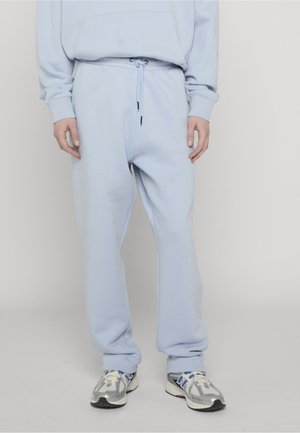 Person trägt hellblaue Jogginghose mit Kordelzug und passendem Sweatshirt, steht mit locker an den Seiten hängenden Händen, weiße und blaue Sportschuhe sichtbar.