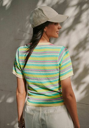 Femeie văzută din profil, purtând o șapcă bej, top cu dungi multicolore și mâneci scurte, pantaloni albi și o brățară, cu umbre pe un perete texturat.