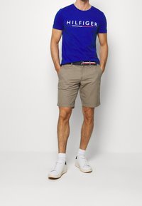 Hombre vestido con una camiseta azul de Hilfiger, pantalones cortos beiges, calcetines blancos, zapatillas blancas y un cinturón negro con un patrón en rojo, blanco y azul.