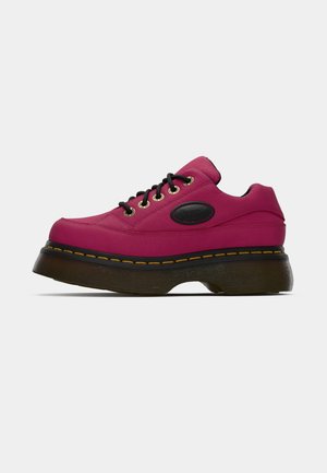 Magenta platform sneaker met zwarte veters, gouden ogen, gele stiksels en dikke doorzichtige bruine zool op een witte achtergrond.