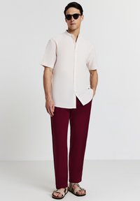 Uomo che indossa una camicia a maniche corte color crema con colletto alla mandarina, pantaloni bordeaux, sandali beige e occhiali da sole scuri, in piedi con una mano in tasca.