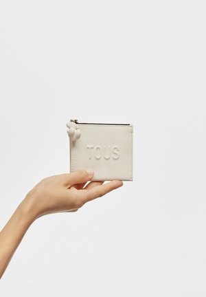 Tarjetero blanco texturizado con el logo "TOUS" en relieve, acentos decorativos redondeados y cierre de cremallera, sostenido en una mano.