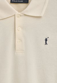 Beżowa koszulka polo z teksturowanej tkaniny, z trzema guzikami przy dekolcie oraz małym logo golfisty w granatowym kolorze na lewej piersi.