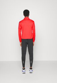Maglia rossa a maniche lunghe con colletto alto, dettagli blu sui polsini, abbinata a pantaloni neri slim con motivo zigzag bianco alle caviglie.