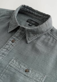 Harmaa napillinen paita, jossa on teksturoitu kangas, terävä kaulus ja yksi rintatasku. Has ruskeat napit ja merkin etiketti.