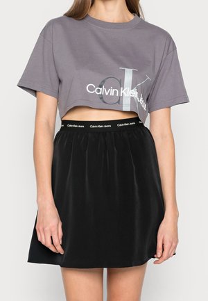 Kobieta ubrana w szarą krótką koszulkę Calvin Klein Jeans oraz czarną spódnicę z elastycznym paskiem z logo Calvin Klein Jeans.