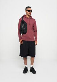 Mørkerød hettegenser, svarte shorts og svarte sneakers. Svart messenger bag med glidelåsdetalj. Modellen har solbriller på seg og står foran en ensfarget bakgrunn.