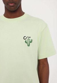 Homme portant un T-shirt vert clair avec un petit cactus vert brodé et le texte noir "Closeo" sur le côté gauche de la poitrine.