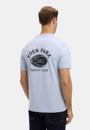 Homme portant un polo à manches courtes bleu clair avec "EDEN PARK", un logo de nœud papillon, et "French Flair" imprimés au dos, tourné de dos.