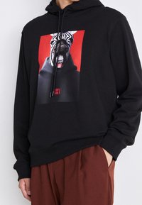 Sweat à capuche noir avec un grand motif de zèbre sur fond rouge. Comprend une capuche à cordon et des poignets côtelés.