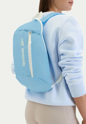 Persona que lleva una sudadera azul claro y pantalones beige con una mochila Reebok azul claro que presenta un bolsillo frontal vertical con cremallera.