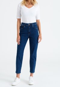 Modré džíny s vysokým pasem a lehkou texturou, pěti kapsami a zapínáním na knoflíky, doplněné těsně padnoucím bíl ým bavlněným tričkem a bílými teniskami.