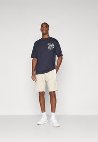 Marine T-skjorte med hvite grafiske detaljer, parret med lysebeige shorts. Modellen har på seg hvite sneakers og ankelstrømper, stående mot en ensfarget bakgrunn.