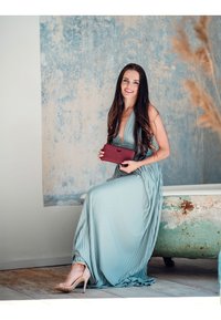 Light blue pleated dress with a deep neckline, holding a burgundy wallet, seated on a vintage, distressed bathtub in a softly lit setting.