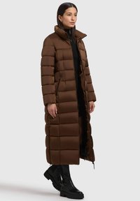 Lang brun pufferjakke med høj krave og quiltet design. Har lynlåsede sidelommer og en glat tekstur. Båret over en sort top og bukser.