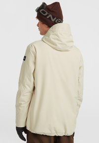 Lichtbeige geïsoleerde jas met capuchon, met een gladde textuur en zwarte accenten. Draag met een bruine muts en zwarte handschoenen.