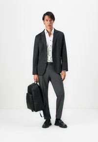 Mann in dunkel kariertem Blazer, weißem Hemd, grauen Hosen, schwarzen Schuhen, der einen schwarzen Leder-Rucksack vor einem weißen Hintergrund hält.