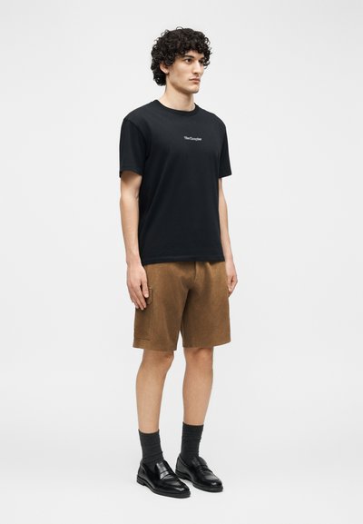 Νέος άνδρας όρθιος, φορώντας μαύρο T-shirt με μικρό κείμενο, καφέ σορτς μέχρι το γόνατο, σκούρες κάλτσες και μαύρα επίσημα παπούτσια, σε απλό φόντο.