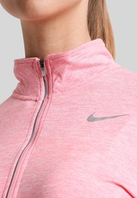 Jasnoróżowa kurtka Nike na zamek błyskawiczny z wysokim kołnierzem, z szarym logo i teksturowanym materiałem, podkreślająca dopasowany krój.