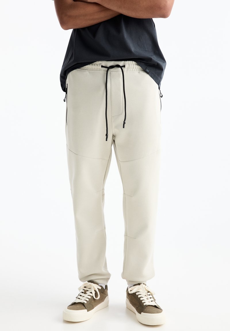 Helle beige Jogginghosen mit elastischem Bund und schwarzen Kordeln, ausgestattet mit Seitentaschen und einem glatten, strukturierten Stoff. Dazu passende braune Sneakers.