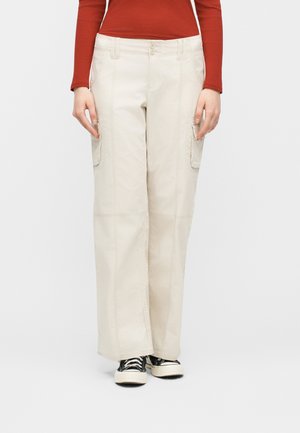 Persona che indossa pantaloni cargo beige a gamba larga con tasche laterali, sneakers bianche e nere e una maglia a maniche lunghe a coste di colore rosso ruggine.